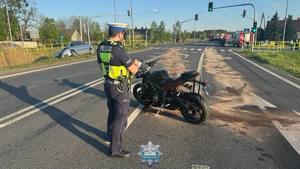 Policjant stoi obok motocykla, który brał udział w zdarzeniu drogowym. Na drodze jest rozsypany sorbent