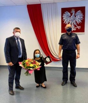 Policjant oraz mężczyzna w garniturze oraz kobieta stojąca z kwiatami i dyplomem
