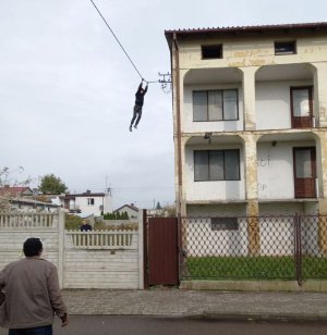 Budynek, mężczyzna wiszący na kablach elektrycznych oraz w lewym dolnym rogu zdjęcia mężczyzna odwrócony tyłem