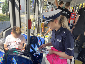 Na zdjęciu widać wnętrze autobusu komunikacji miejskiej. Wśród pasażerów znajduje się policjant w granatowym mundurze i białej czapce z napisem „POLICJA”. Funkcjonariusz stoi obok siedzącego dziecka, którego twarz została zasłonięta pikselami. Policjant trzyma w ręku materiały edukacyjne oraz różowy worek. W tle widać innych pasażerów siedzących i stojących w autobusie.