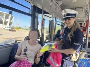 Zdjęcie przedstawia wnętrze autobusu. Policjantka w granatowym mundurze i białej czapce wręcza siedzącemu pasażerowi elementy odblaskowe oraz różowy worek.