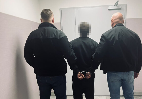 Zatrzymany mężczyzna, ktory ma założone na ręce trzymane z tyłu kajdanki. Prowadzi go dwóch policjantów po cywilnemu.