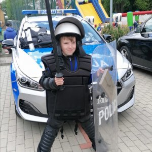 Dziecko ubrane w wyposażenie policyjne z tarczą - przy radiowozie oznakowanym