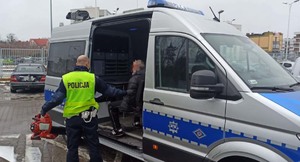 Na zdjęciu policjant umundurowany trzyma w ręku odzyskany po kradzieży sprzęt ogrodniczy, w tle podejrzany siedzi w radiowozie