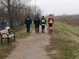 Na zdjęciu tyłem ( policjant, strażak oraz ratownik medyczny) idą w rejonie wału nad rzeką Narew i kontrolują miejsca, w których mogą przebywać osoby bezdomne, aby uchronić ich przed wychłodzeniem organizmu