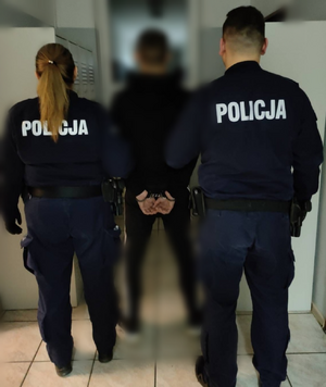 policjanci stoją tyłem, pomiędzy nimi zatrzymany mężczyzna stoi w kajdankach