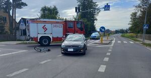 Miejsce zdarzenia drogowego, na zdjęciu opel, rower leżący na drodze, w tle radiowóz i pojazd straży pożarnej