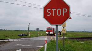 Zdjęcie przedstawiające znak drogowy ,,Stop" na przejeździe  kolejowym, w tle widoczny rozbity samochód i auto policji i strazy pożarnej