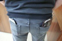 Zdjęcie przedstawia mężczyznę stojącego tyłem, w kieszeniach jego spodni znajduje się portfel i telefon