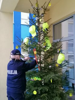 Na zdjęciu widzimy umundurowanego policjantkę, która wiesza elementy odblaskowe na choinkę