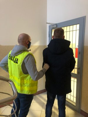 Na zdjęciu widać policjanta podczas wykonywania czyności z osobą zatrzymaną, w tle wejście do pomieszczenia dla osób zatrzymanych