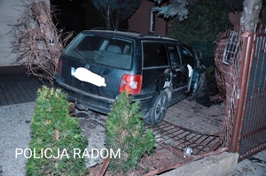 Zdjęcie przedstawia samochód marki volkswagen uszkodzony z przodu i prawej strony. Znajdujący się na prywatnej posesji, wraz z pojazdem zostało uszkodzone ogrodzenie posesji.