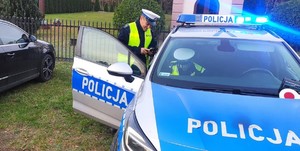 Radiowóz, a w nim jeden z policjantów z drogówki. Drugi umundurowany policjant jest obok auta.