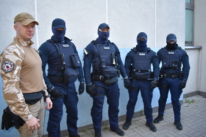 Na zdjęciu jest 5 umundurowanych policjantów. czterech z nich jest w granatowych mundurach i maja zasłonięte twarze, natomiast po lewej stronie jest policjant w jasnym mundurze i ma czapkę