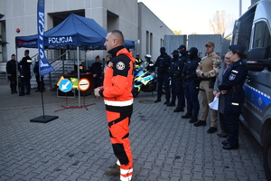 Na środku stoi policyjny ratownik. W tle grupa mundurowych i namiot z napisem Policja.