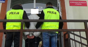 Dwóch policjantów w żółtych odblaskowych kamizelkach z napisem policja prowadzi zatrzymanego mężczyznę. 
Zatrzymany ma założone na ręce kajdanki trzymane z tyłu.