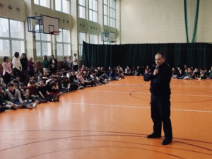 dzielnicowy na sali gimnastycznej z dziećmi podczas spotkania, policjant trzyma mikrofon, po lewej stronie siedzą uczniowie