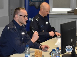 dwaj policjanci siedzą przy stole, z lewej strony mężczyzna trzyma mikrofon i jest w okularach