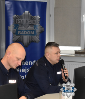 dwaj policjanci w mundurach, jeden  z nich trzyma mikrofon a w tle baner z napisem KMP w Radomiu