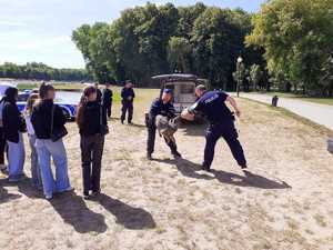 uczniowie na plaży nad zalewem obserwują policyjny pokaz z udziałem psa służbowego, w tle radiowóz