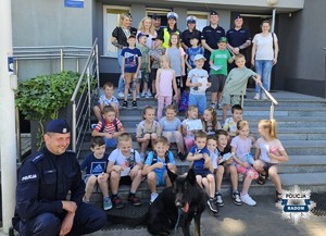 zdjęcie grupowe, dzieci, policjanci i opiekunowie na schodach przed budynkiem komisariatu, po lewej stronie policjant  z psem słuzbowym