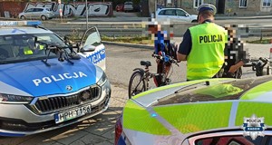 dwa radiowozy w srodku tyłem stoi policjant kontroluje 2 rowerzystów