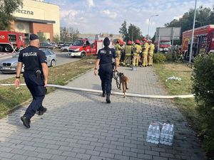 policjant i policjantka z psem służbowym idą chodnikiem w tle grupa strażaków