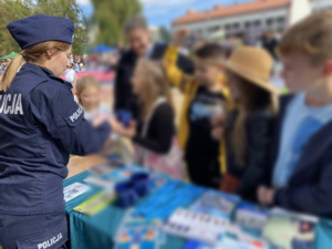 Policjantka tyłem przy stoliku profilaktycznym wręczająca dzieciom gadżety z logo policja. Twarze dzieci rozmazane