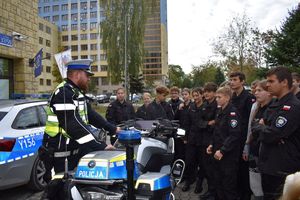 policjant z drogówki stoi przy policyjnym motocyklu za nim radiowóz po prawej stronie uczniowie klasy policyjnej