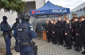 po lewej stronie trzech policjantów z grupy realizacyjnej mają zasłonięte twarze po prawej stronie uczniowie w tle namiot z napisem policja