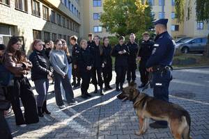 po prawej stronie przed budynkiem komendy stoi policjant obok policyjny pies, po lewej grupa uczniów