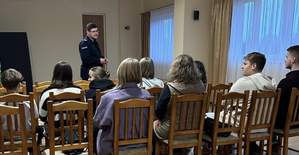 policjant prowadzący spotkanie, przodem do aparatu. na ekranie widoczni uczestnicy siedzący na krzesłach tyłem do aparatu.