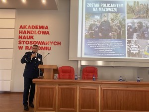 policjant na sali wykładowej trzyma mikrofon , za nim ekran z prezentacja napis na niej zostań policjantem na mazowszu, w tle na ścianie napis Akademia Handlowa Nauk Stosowanych w Radomiu