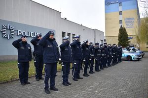 grupa policjantów stoi w dwuszeregu po prawej w tle radiowozy i budynek komendy