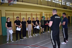 policjanci w strojach sportowych tyłem do aparatu w tle grupa uczniów na sali gimastycznej