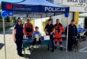 pod niebieskim namiotem stoisko profilaktyczne, jest troje policjantów, dziecko policyjny pies dwóch męzczyzn w czerwonych strojach