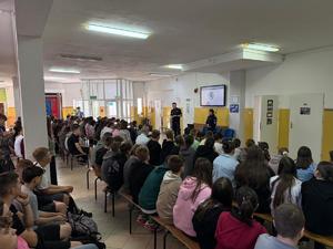 w szkole na ławkach siedzi duża grupa uczniów w tle policjant i policjantka oraz ekran