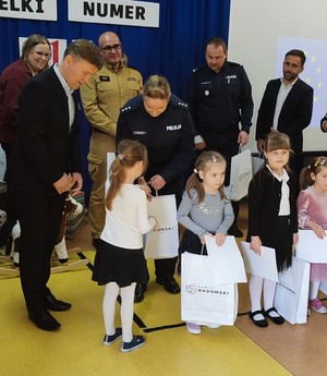 policjantka wrecza upominek dziecku dookoła dzieci, mundurowi po lewej mężczyzna w garniturze