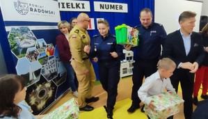 policjantka i policjant ida z upominkiem do dziecka laureata konkursu, w tle mundurowy i męzczyzna w garniturze