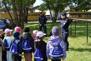 grupa dzieci z niebieskimi plecakami w tle dwoje policjantów i pies słuzbowy