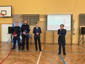 na sali gimnastycznej policjanci i dyrektor edukacji na srodku sali policjantka trzyma mikrofon
