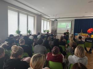 spotkanie na sali z młodzieżą uczestników widać tyłem, siedzących na krzesłach w tle kobieta psycholog za nia ekran