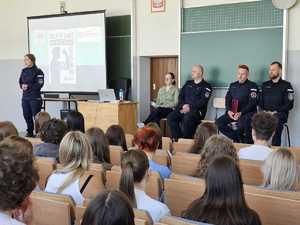 uczniowie siedza w ławkach widac ich tylem w tle policjanci i ekran