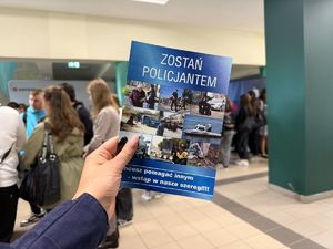 dłoń policjantki z ulotka z napisem zostań policjantem w tle grupa studentów