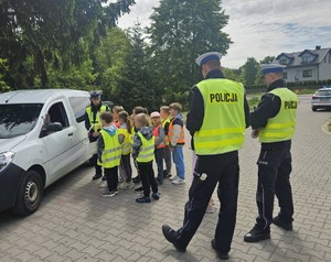 policjanci i grupa dzieci w kamizelkach odblaskowych stoją przy białym aucie