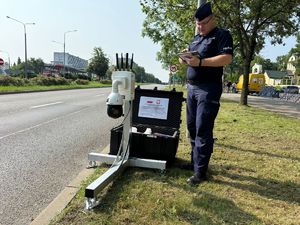 Umundurowany policjant stoi obok drogi, trzyma tablet. Obok policjanta znajduje się kamera Obserwator City.