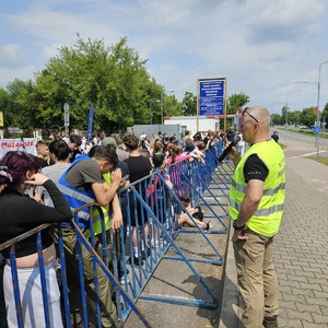 Mężczyzna w odblaskowej kamizelce stoi z mikrofonem przy barierach ochronnych. Po drugiej stronie barier na parkingu znajdują się dzieci.