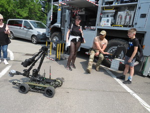 Stoisko policyjne z robotem pirotechnicznym- demonstracja sprzętu policyjnego. Policjant prezentuje sprzęt, trzy osoby uważnie słuchają.