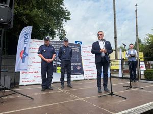 Trzech mężczyzn stoi na scenie. Dwóch umundurowanych policjantów stoi z tyłu natomiast trzeci w ciemnej marynarce i białej koszuli, mówi do mikrofonu.
