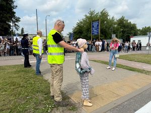 Mężczyzna w odblaskowej kamizelce trzyma manekina obok przejścia dla pieszych w tle znajdują się obserwatorzy, goście wydarzania.